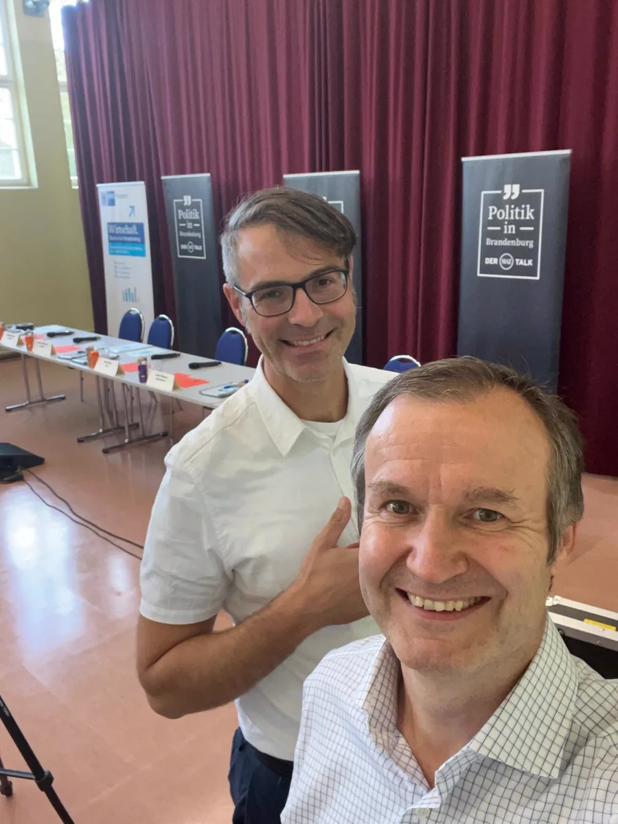 Zwei Mitglieder der Wirtschaftsjunioren vor der Bühne einer politischen Gesprächsrunde, im Hintergrund Roll-ups und die vorbereitete Podiumstafel.
