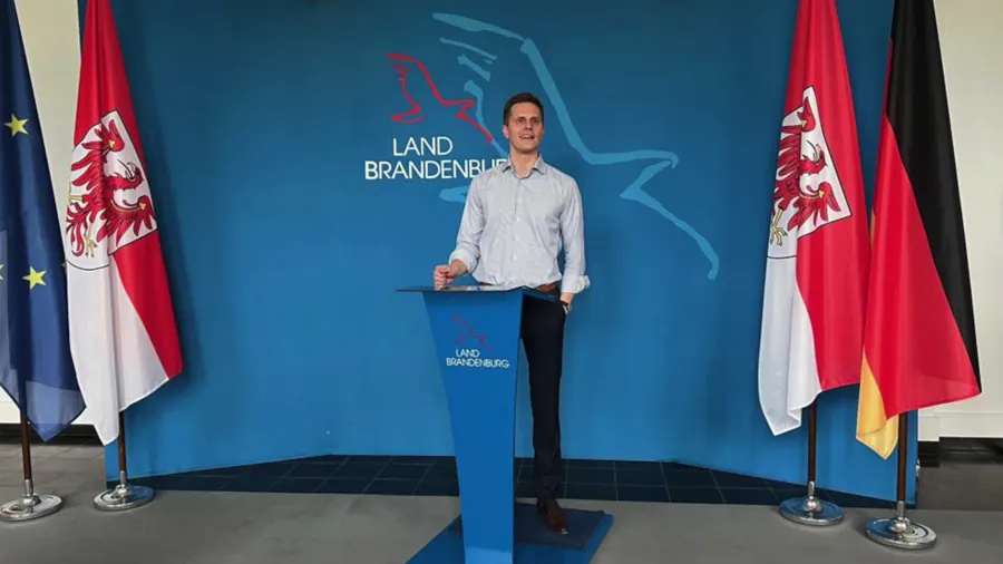 Person an einem Rednerpult mit dem Wappen des Landes Brandenburg, dahinter die Flaggen von Europa, Brandenburg und Deutschland.