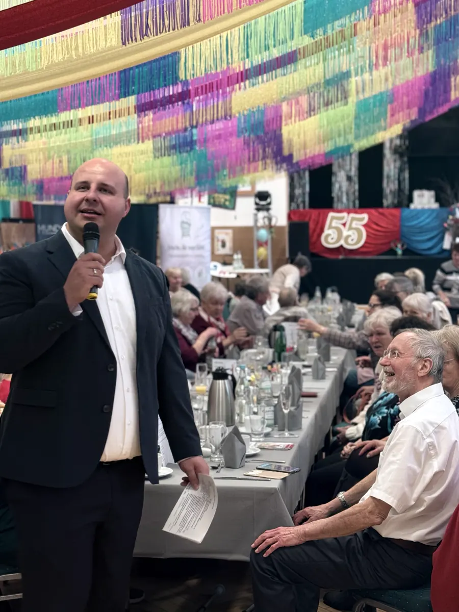 Moderator mit Mikrofon vor langen Tischen mit älteren Gästen, darüber bunte Girlanden.